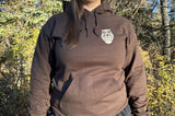 Person wearing a brown hoodie with a cream owl logo in a forest setting