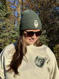 Person wearing a green beanie and sunglasses with a logo on a beige sweatshirt in a forest setting